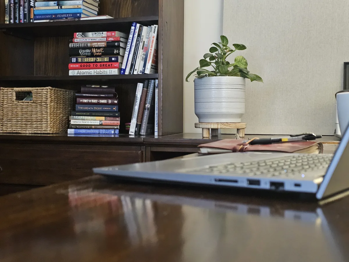 A bookshelf of leadership books with a laptop and plant on a wood desk