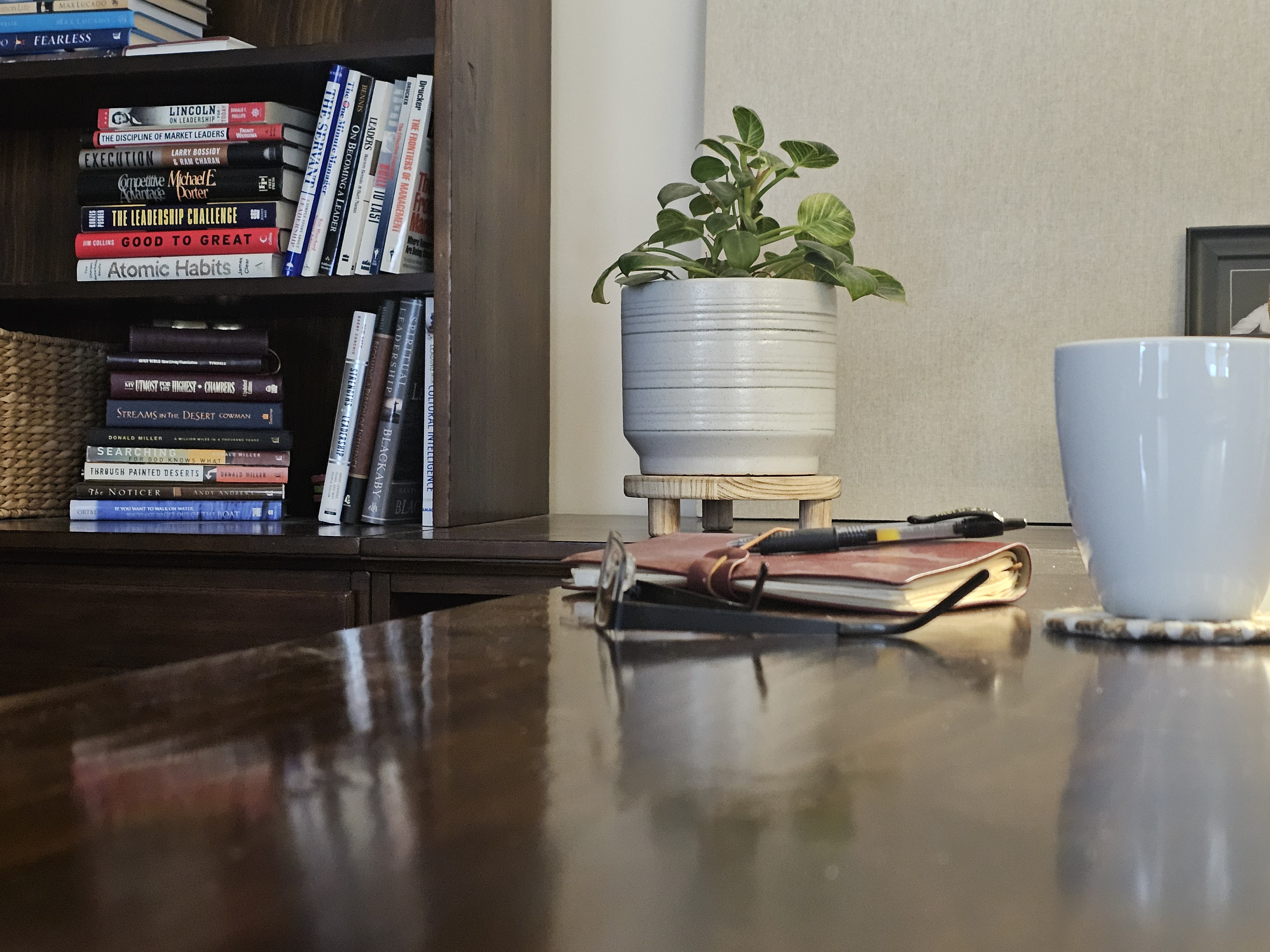 A bookshelf with a journal, pen, plant, and coffee mug on a wood desk