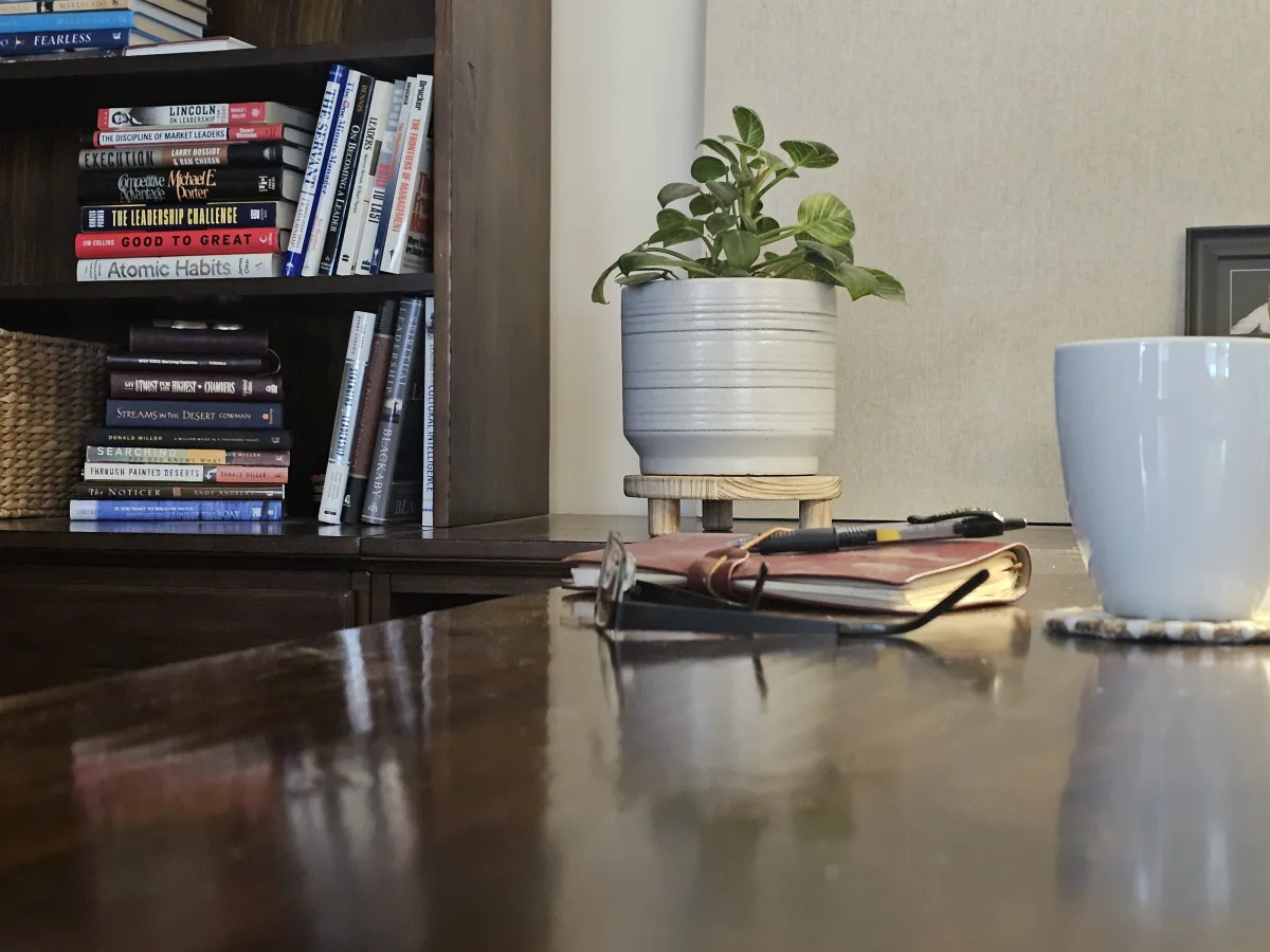 A bookshelf with a journal, pen, plant, and coffee mug on a wood desk